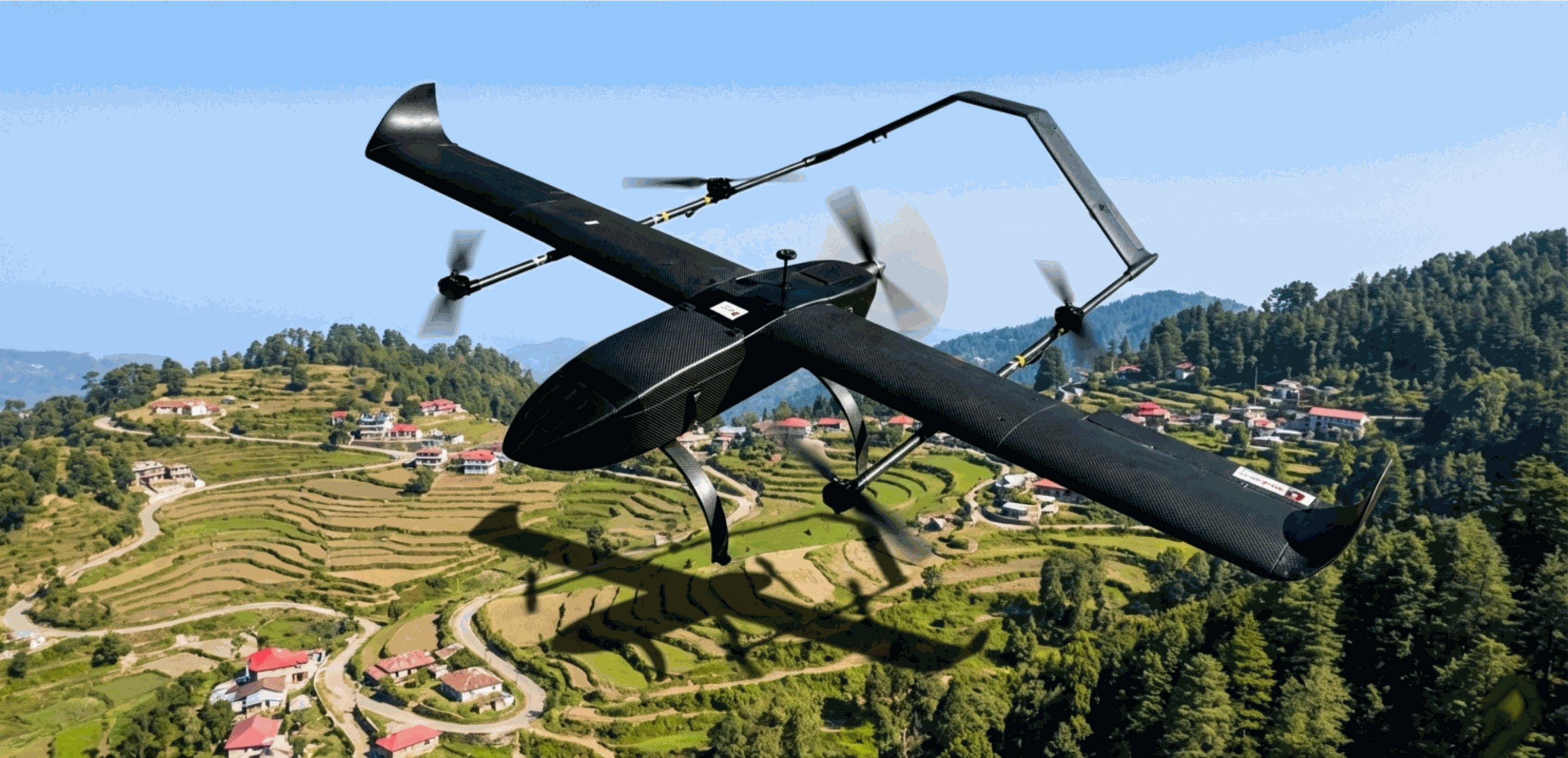 Hans Isometric view of black Hans VTOL fixed-wing drone flying over green terraced fields and a hill station village. The drone casts a shadow on the ground, displaying its hybrid vertical lift and forward flight propellers.VTOL hybrid drone conducting precision aerial survey over complex hilly terrain. Designed for high-altitude mapping and long-range surveillance in India's toughest landscapes.