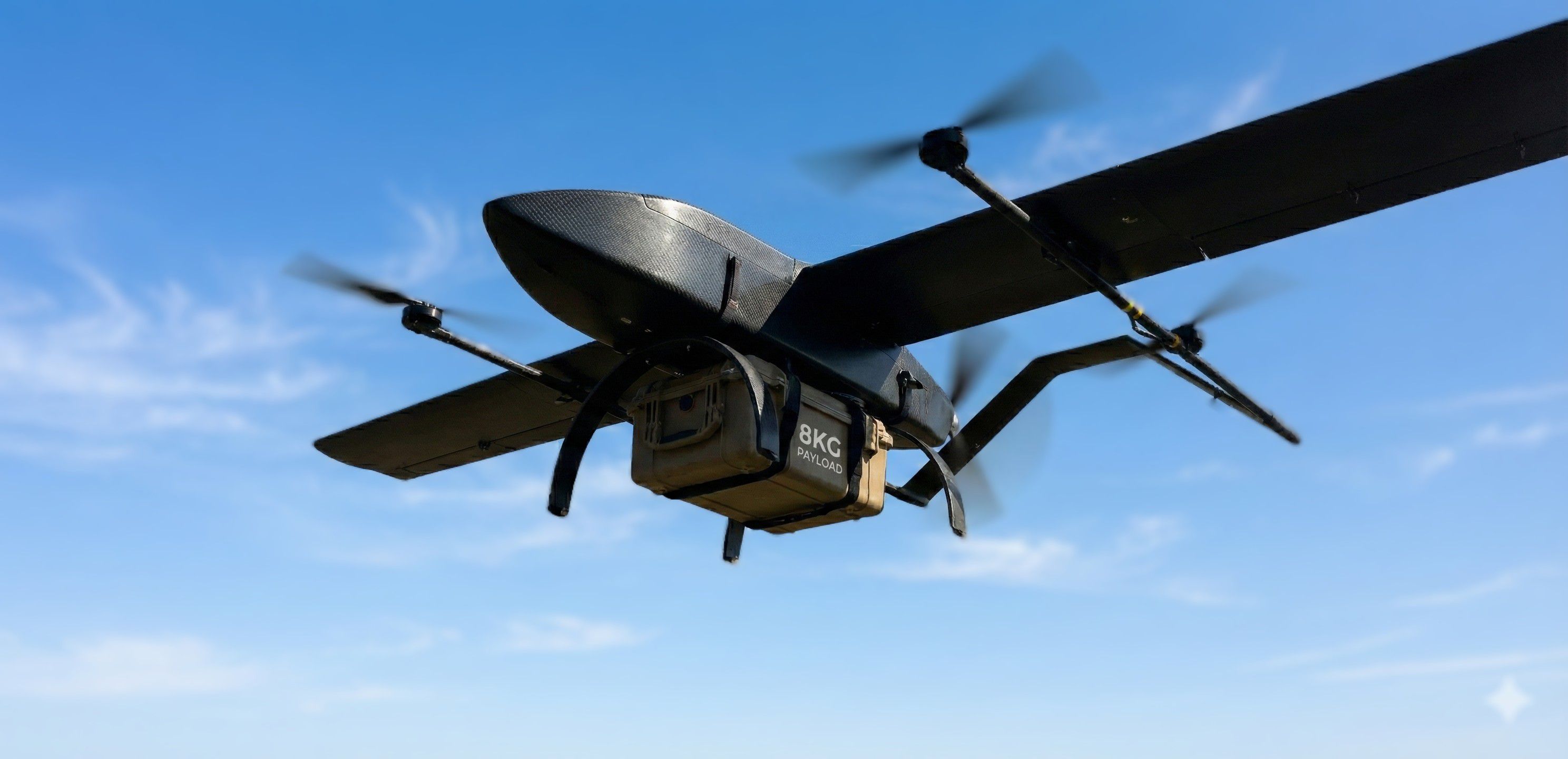 Low-angle view of a black Hans VTOL drone flying against a blue sky, carrying a beige rugged cargo case labeled '8KG PAYLOAD' mounted to its underbelly.